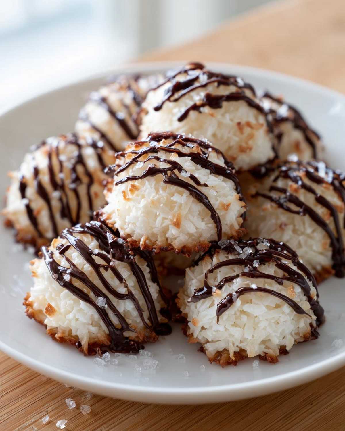A pile of homemade Coconut Macaroons with Dark Chocolate Drizzle, sprinkled with sea salt, served on a white plate.
