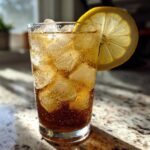 A tall glass filled with ice and dark brown Cold Brew Lemonade, garnished with a bright lemon slice.