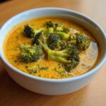 Close-up of a white bowl filled with vibrant orange Copycat Broccoli Cheddar Soup, topped with large florets of bright green broccoli.