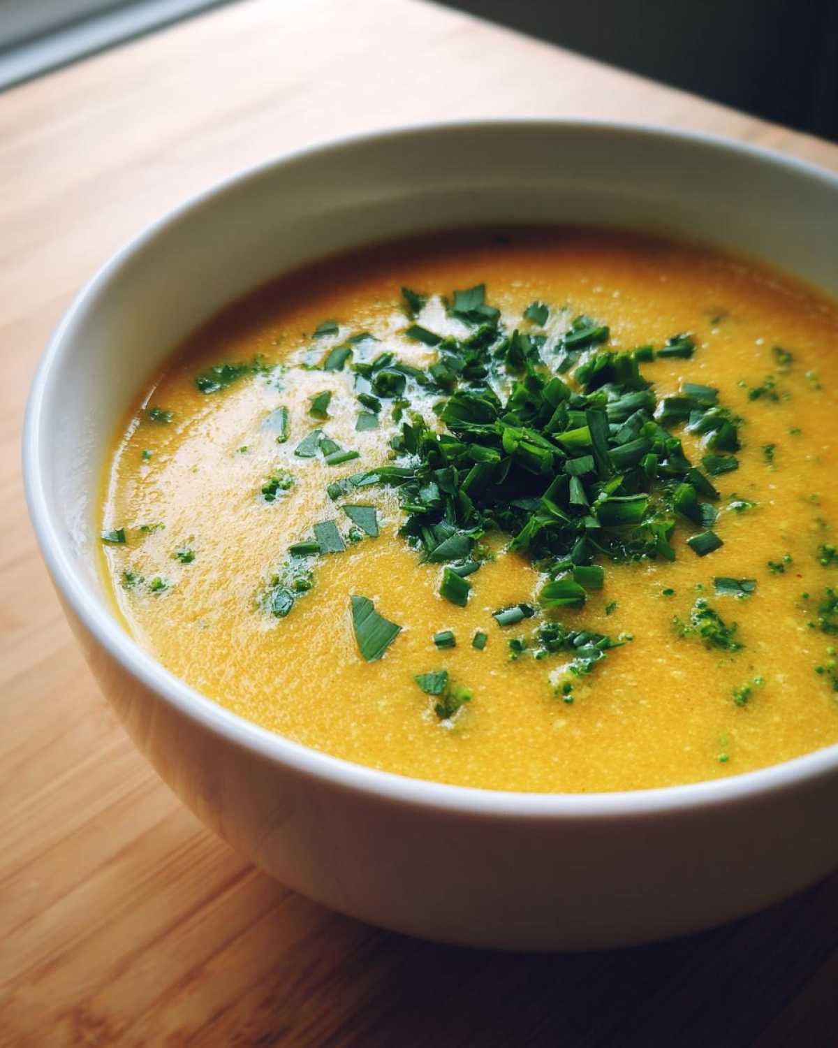 Close-up of a creamy, bright yellow bowl of Copycat Broccoli Cheddar Soup topped generously with fresh, chopped green chives.