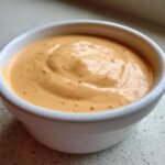 A close-up, featured image of creamy, orange-hued Copycat Raising Cane’s Sauce speckled with red spices, served in a small white ramekin.
