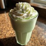 A tall glass filled with a light green Copycat Shamrock Shake topped with a swirl of white mint whipped cream.