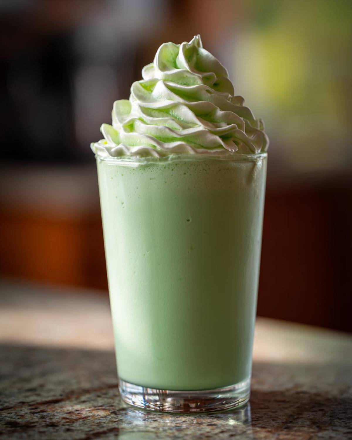 A tall glass filled with a thick, light green Copycat Shamrock Shake topped with a swirl of pale green Mint Whipped Cream.