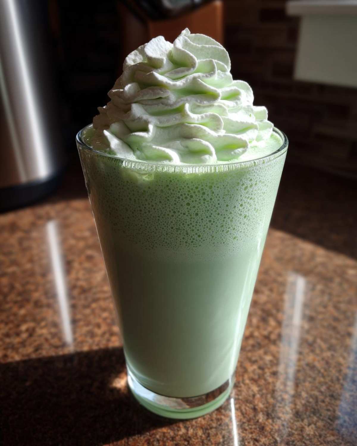 A tall glass filled with a thick, green Copycat Shamrock Shake topped with light green mint whipped cream.
