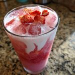 A tall glass filled with the pink and white layers of a Copycat Starbucks Pink Drink, topped with ice and sliced strawberries.