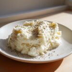 A mound of fluffy Creamy Garlic Mashed Cauliflower topped with cracked black pepper and herbs on a white plate.