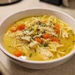 Close-up of a white bowl filled with Creamy Lemon Chicken Orzo Soup, topped with shredded chicken, carrots, and parsley.