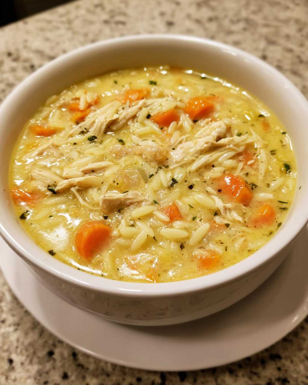Close-up of a white bowl filled with Creamy Lemon Chicken Orzo Soup, featuring shredded chicken, orzo pasta, and bright orange carrots.