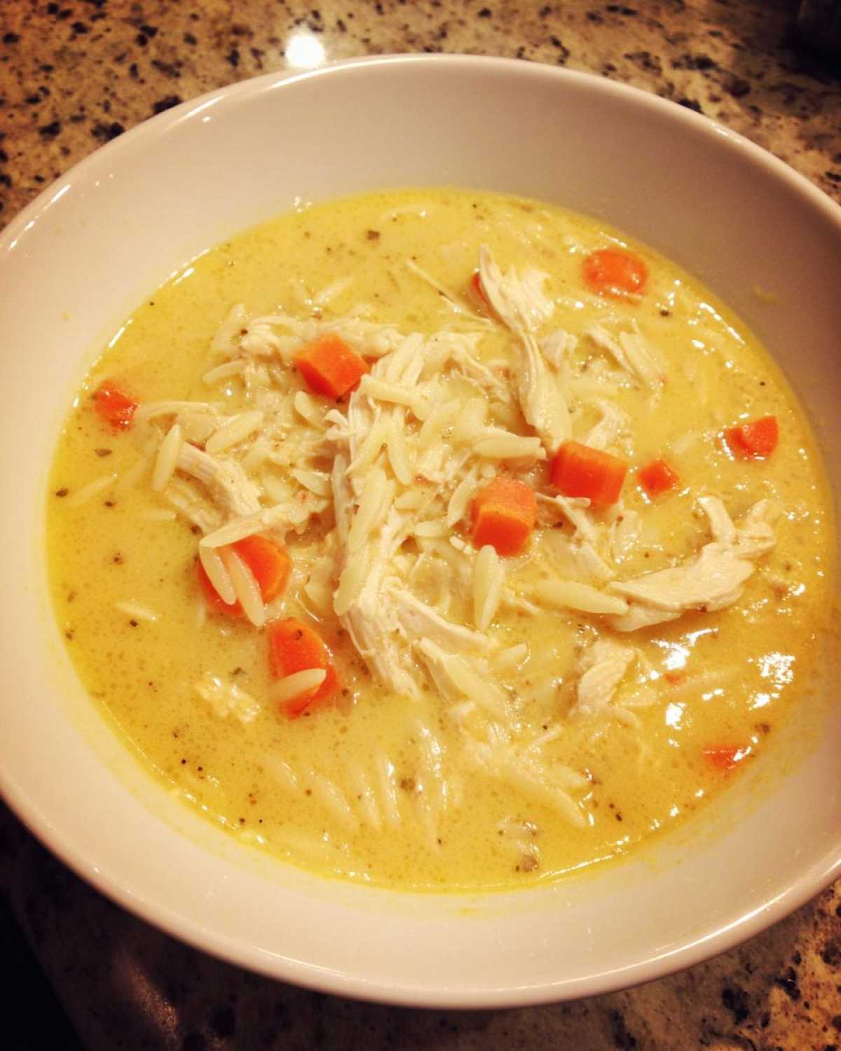 A close-up of a white bowl filled with Creamy Lemon Chicken Orzo Soup, featuring shredded chicken, orzo pasta, and diced carrots.