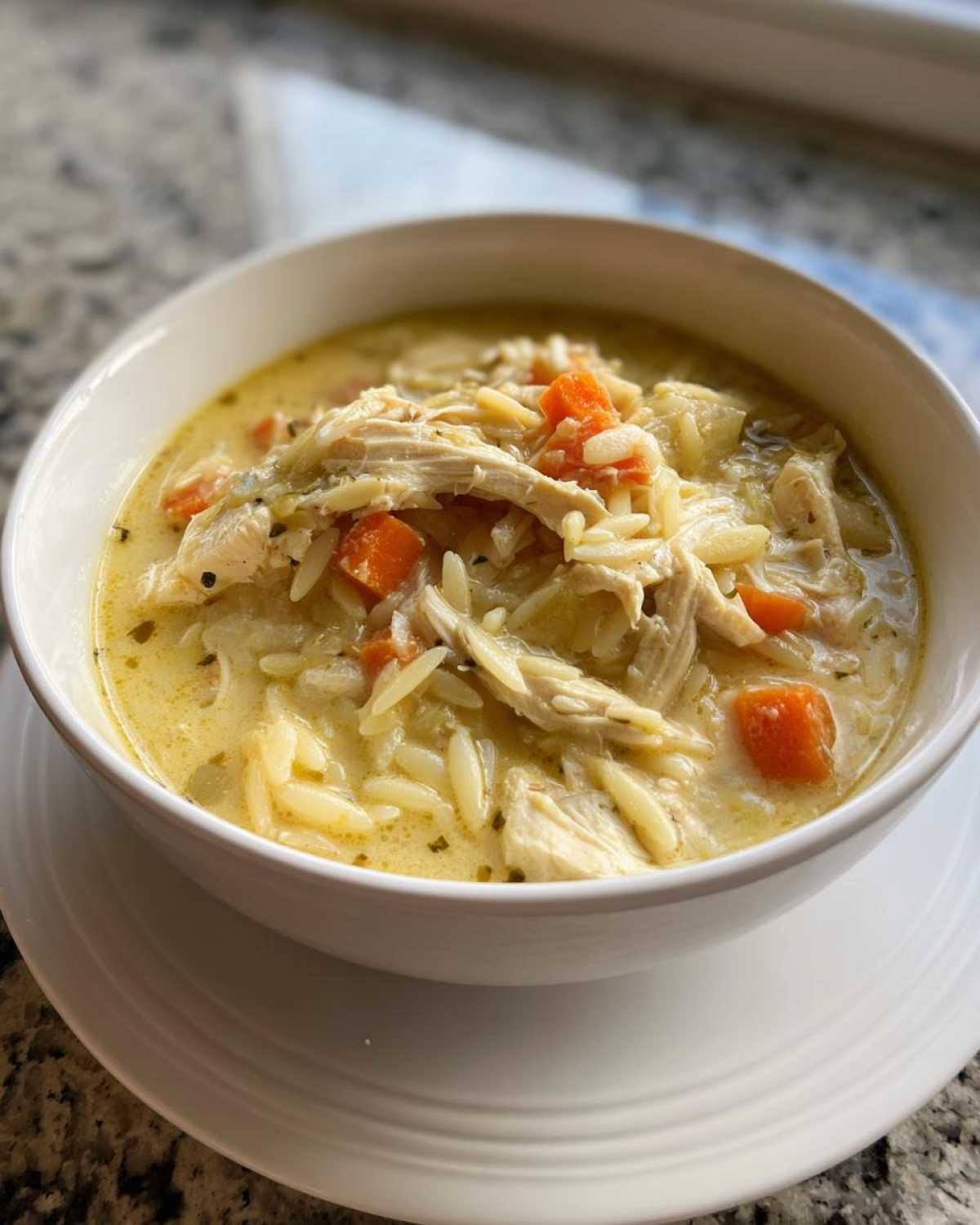 A close-up of a white bowl filled with rich Creamy Lemon Chicken Orzo Soup, featuring shredded chicken, orzo pasta, and diced carrots.