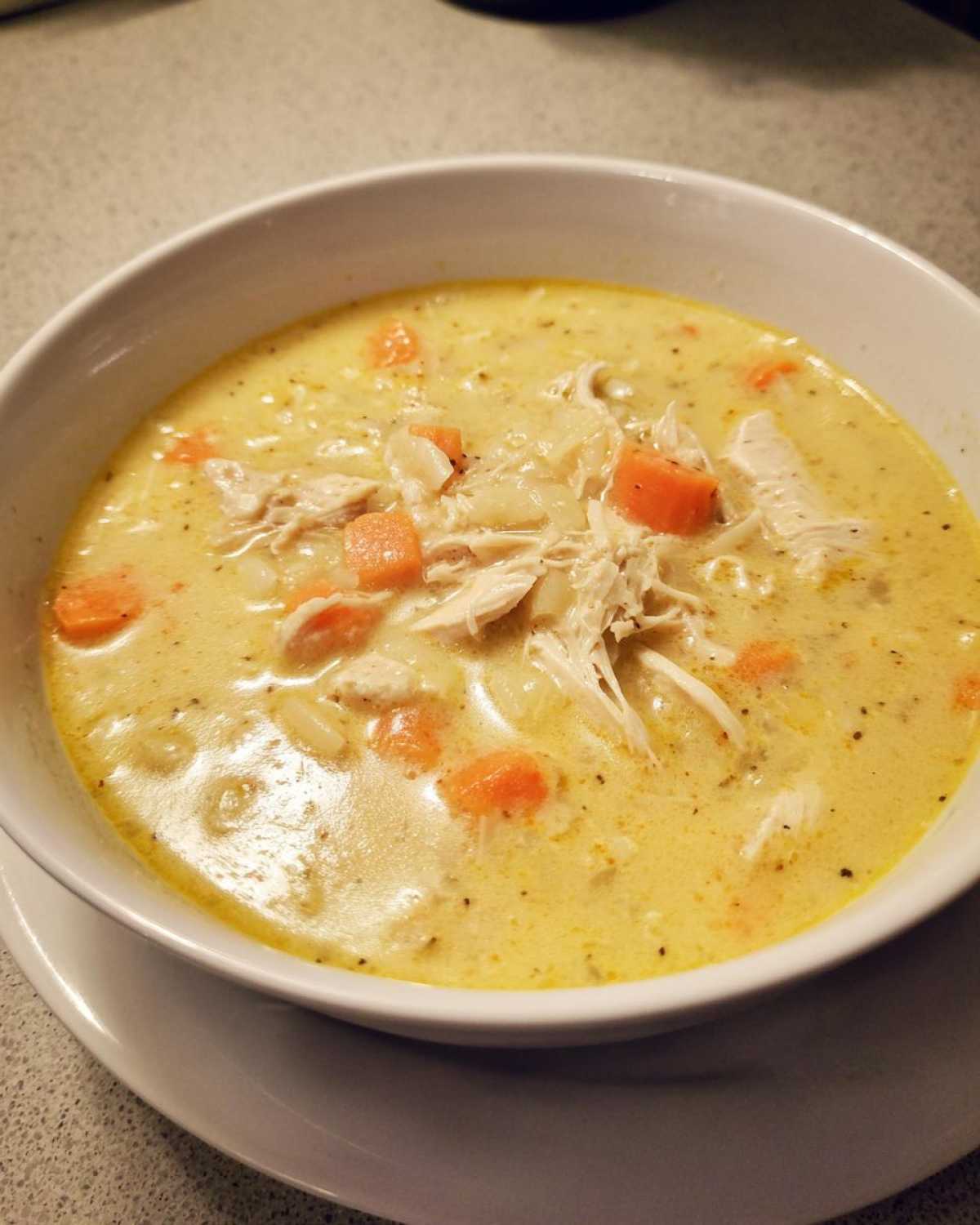 A close-up of a white bowl filled with rich, yellow Creamy Lemon Chicken Orzo Soup, featuring shredded chicken and diced carrots.