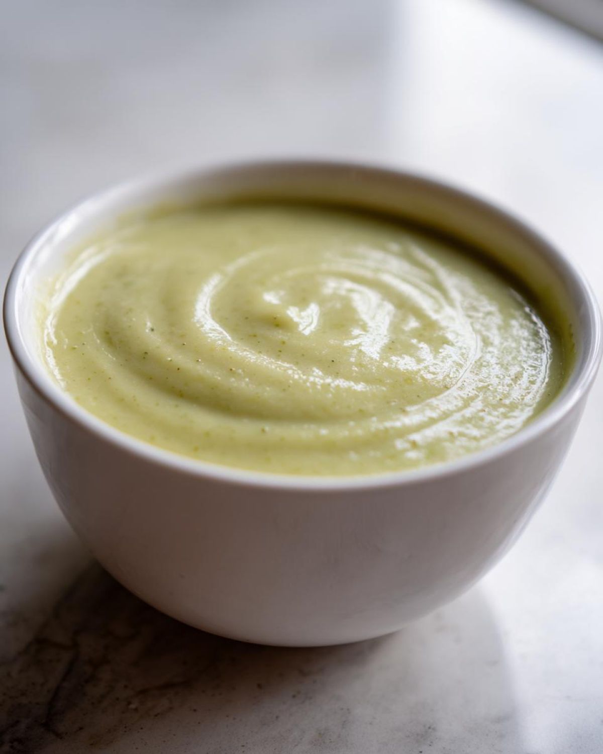 Close-up of a white bowl filled with smooth, pale green Creamy Potato Leek Soup, showing a swirl pattern on top.