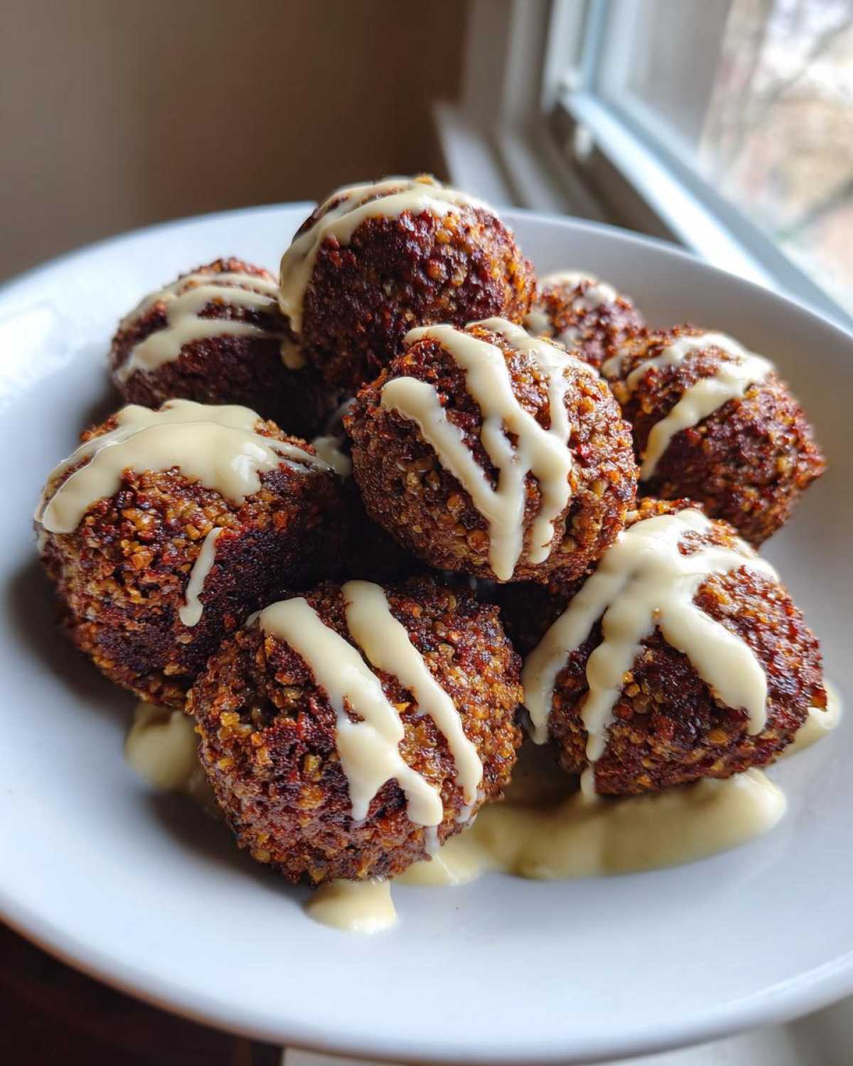 A close-up of several golden-brown, crispy air fryer falafel balls stacked in a white bowl, drizzled generously with creamy tahini sauce.