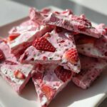 A pile of broken pieces of pink and white Frozen Strawberry Yogurt Bark, studded with sliced strawberries and chia seeds, served on a white plate.