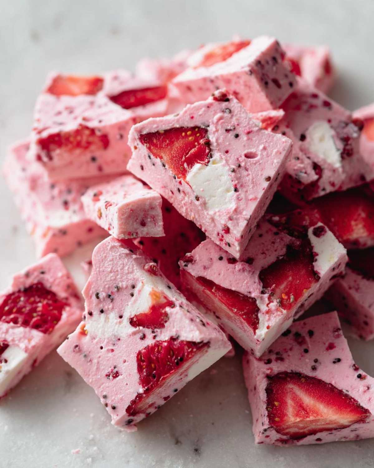 Close-up of stacked, pink, square pieces of Frozen Strawberry Yogurt Bark featuring visible strawberry slices and black chia seeds.