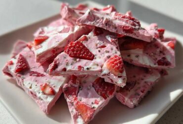 A pile of broken pieces of pink and white Frozen Strawberry Yogurt Bark, studded with sliced strawberries and chia seeds, served on a white plate.