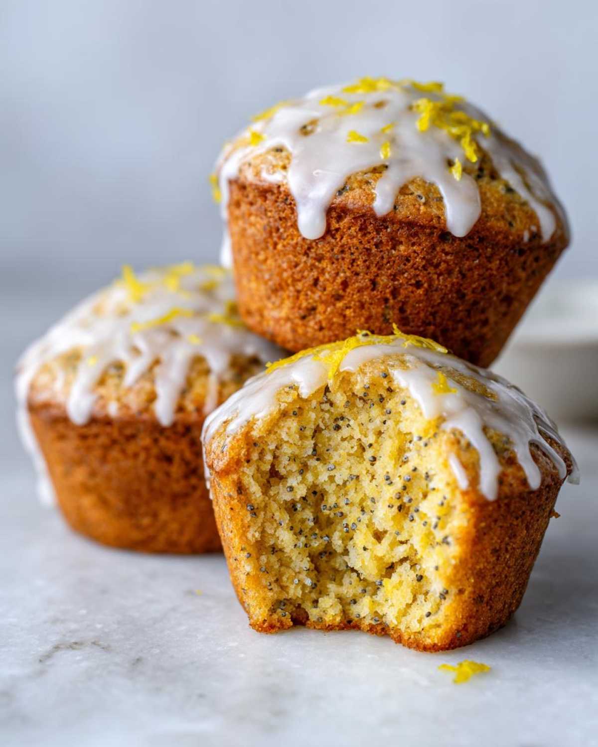 Three Glazed Lemon Poppy Seed Muffins stacked, with one in front showing a bite taken out revealing the moist interior.