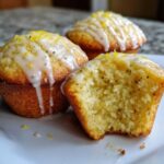 Three Glazed Lemon Poppy Seed Muffins on a white plate, one with a bite taken out showing the moist interior.