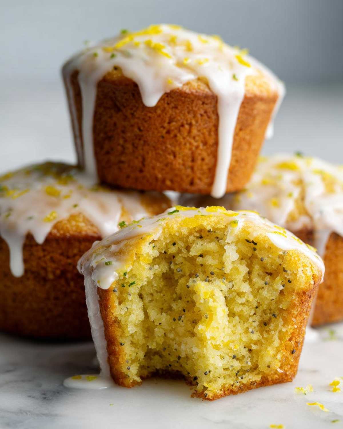 Close-up of Glazed Lemon Poppy Seed Muffins, one with a bite showing the moist, yellow crumb and poppy seeds.