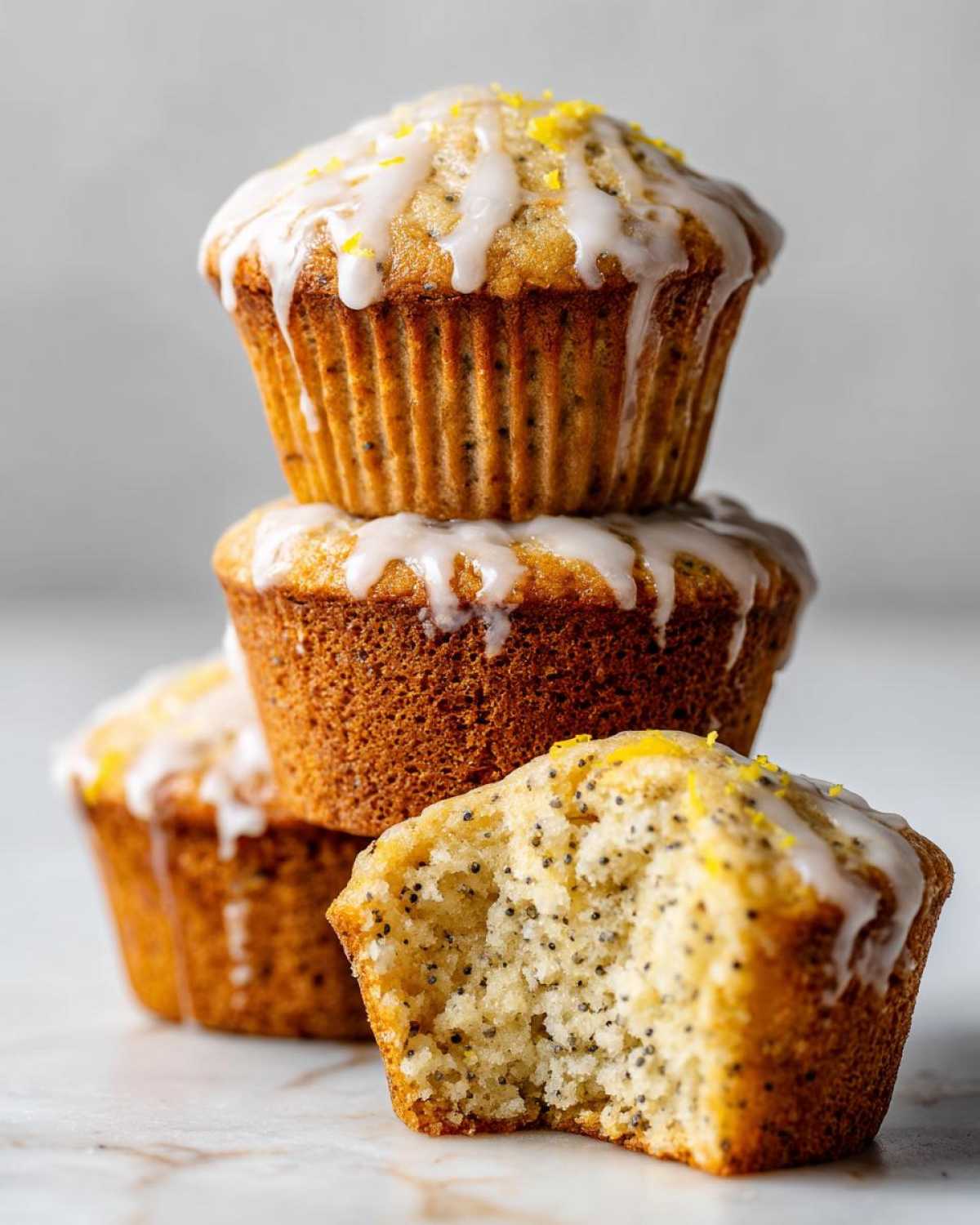 A stack of three Glazed Lemon Poppy Seed Muffins, with one in front having a bite taken out showing the moist crumb and poppy seeds.