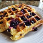 Two golden-brown Gluten-Free Blueberry Waffles loaded with melted, juicy blueberries served on a white plate.
