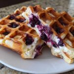 Two halves of golden brown Gluten-Free Blueberry Waffles, showing melted blueberries inside, served on a white plate.