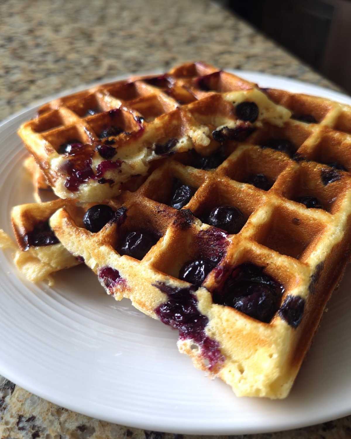 Close-up of golden Gluten-Free Blueberry Waffles stacked on a white plate, featuring burst blueberries.