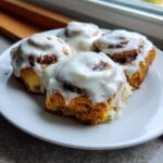 Four soft Gluten-Free Cinnamon Rolls generously topped with white cream cheese frosting on a white plate.