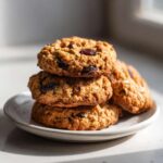 A stack of four golden-brown Gluten-Free Oatmeal Raisin Cookies dotted with dark raisins, resting on a small white plate.