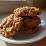A stack of four golden brown Gluten-Free Oatmeal Raisin Cookies resting on a small white plate, highlighted by sunlight.