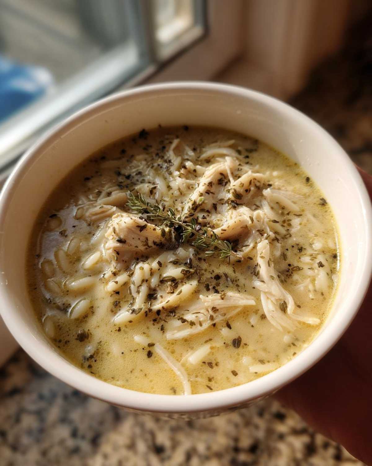 Close-up of a white bowl filled with creamy Greek Lemon Chicken Soup, featuring shredded chicken, orzo pasta, and herbs.