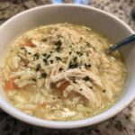 Close-up of a white bowl filled with creamy Greek Lemon Chicken Soup, featuring shredded chicken, orzo pasta, and parsley.