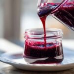 Close-up of thick, rich Homemade Blueberry Syrup being poured from a glass pitcher into a small jar.