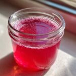 Close-up of bright pink Homemade Strawberry Lemonade Concentrate in a glass jar, catching sunlight.