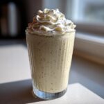 A tall glass filled with a speckled Homemade Vanilla Bean Frappuccino topped high with whipped cream.