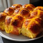 A plate filled with freshly baked, golden brown Hot Cross Buns with Orange Glaze, glistening under natural light.