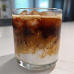 Close-up of a condensation-covered glass containing an Iced Brown Sugar Oatmilk Latte with swirling layers of coffee and oatmilk.
