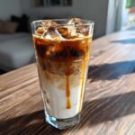 A tall glass of Iced Caramel Macchiato layered with milk, espresso, ice, and dripping caramel sauce.