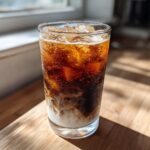 A tall glass showing the layers of an Iced Honey Almond Milk Latte with ice cubes, dark coffee, and swirling almond milk.
