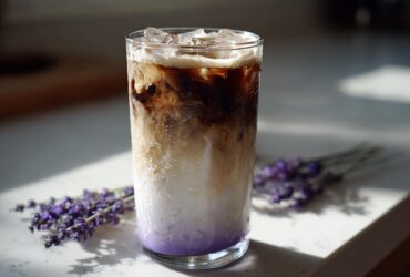 A tall glass of Iced Lavender Latte showing distinct layers of purple syrup, milk, and dark espresso, garnished with ice.