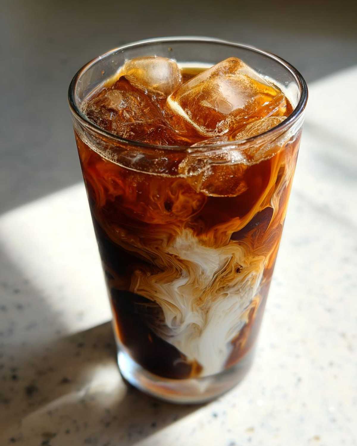 Close-up of a tall glass containing an Iced Mocha, showing rich dark coffee swirling dramatically with white cream over ice cubes.