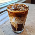 Close-up of a tall glass containing an Iced Mocha with ice cubes, showing rich dark coffee swirling beautifully with white cream.