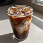 Close-up of a tall glass of Iced Mocha with ice cubes and cream swirling into the dark coffee.
