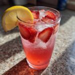 Close-up of a tall glass filled with Iced Strawberry Lemon Tea, ice, sliced strawberries, and a lemon wedge garnish.