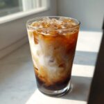 A tall glass filled with an Iced Vanilla Chai Latte showing rich, swirling patterns of dark chai concentrate and white milk over ice.