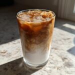 A tall glass of Iced Vanilla Chai Latte showing rich brown chai swirling into white milk over ice cubes.