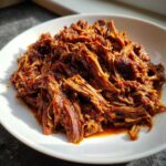 A close-up of saucy, shredded Instant Pot Beef Barbacoa piled high on a white plate.