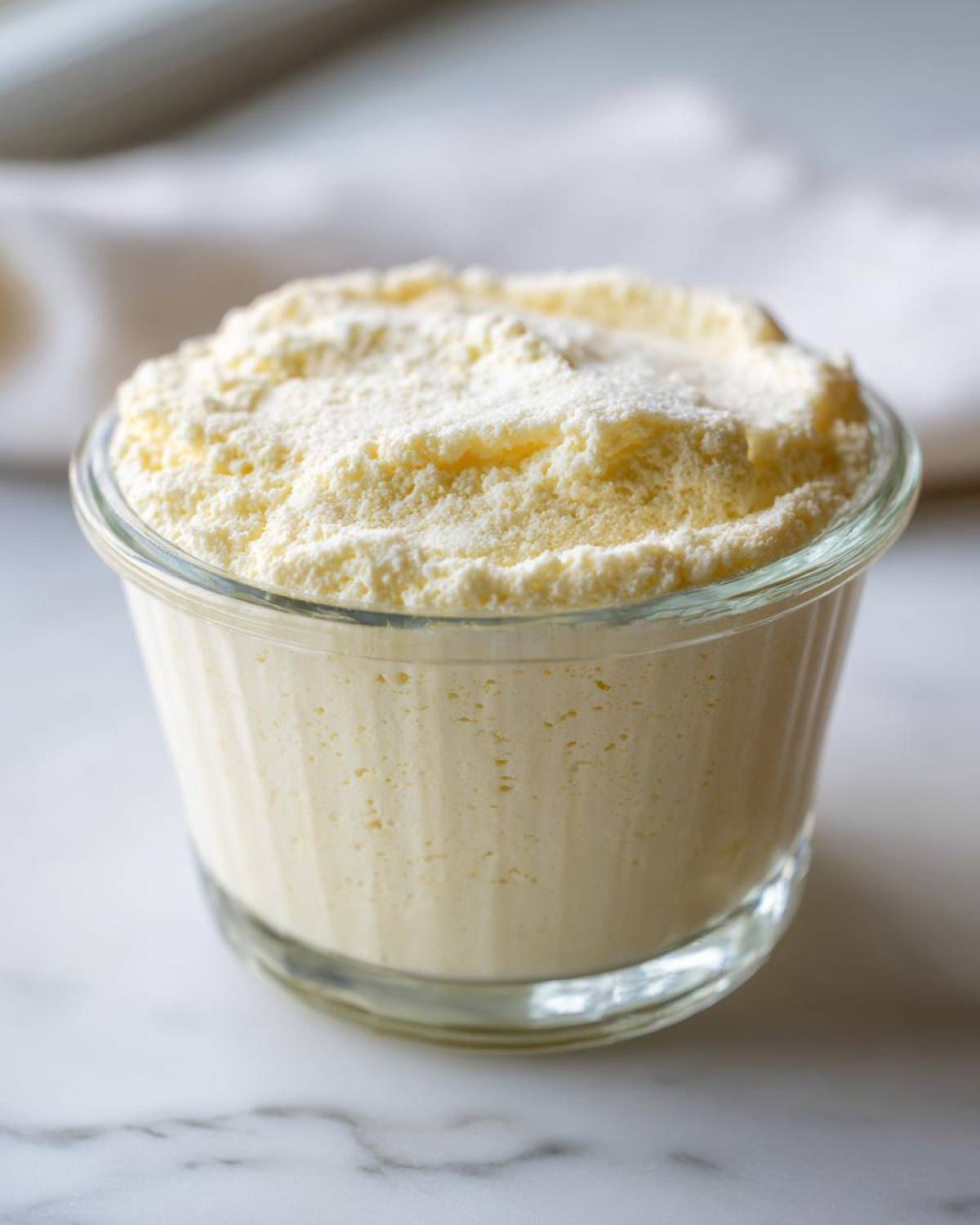 Close-up of a single serving of light yellow Keto Lemon Cheesecake Mousse topped with fine powder in a small glass ramekin.