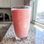 A tall glass filled with a pink, blended Keto Strawberry Cheesecake Smoothie sitting on a granite countertop near a window.