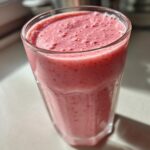 Close-up of a tall glass filled with a thick, pink Keto Strawberry Cheesecake Smoothie, catching bright sunlight.